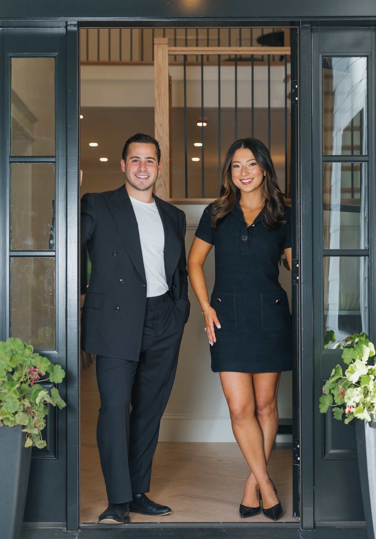 Two real estate professionals pose in a doorway, with a staircase behind them and potted flowers nearby.