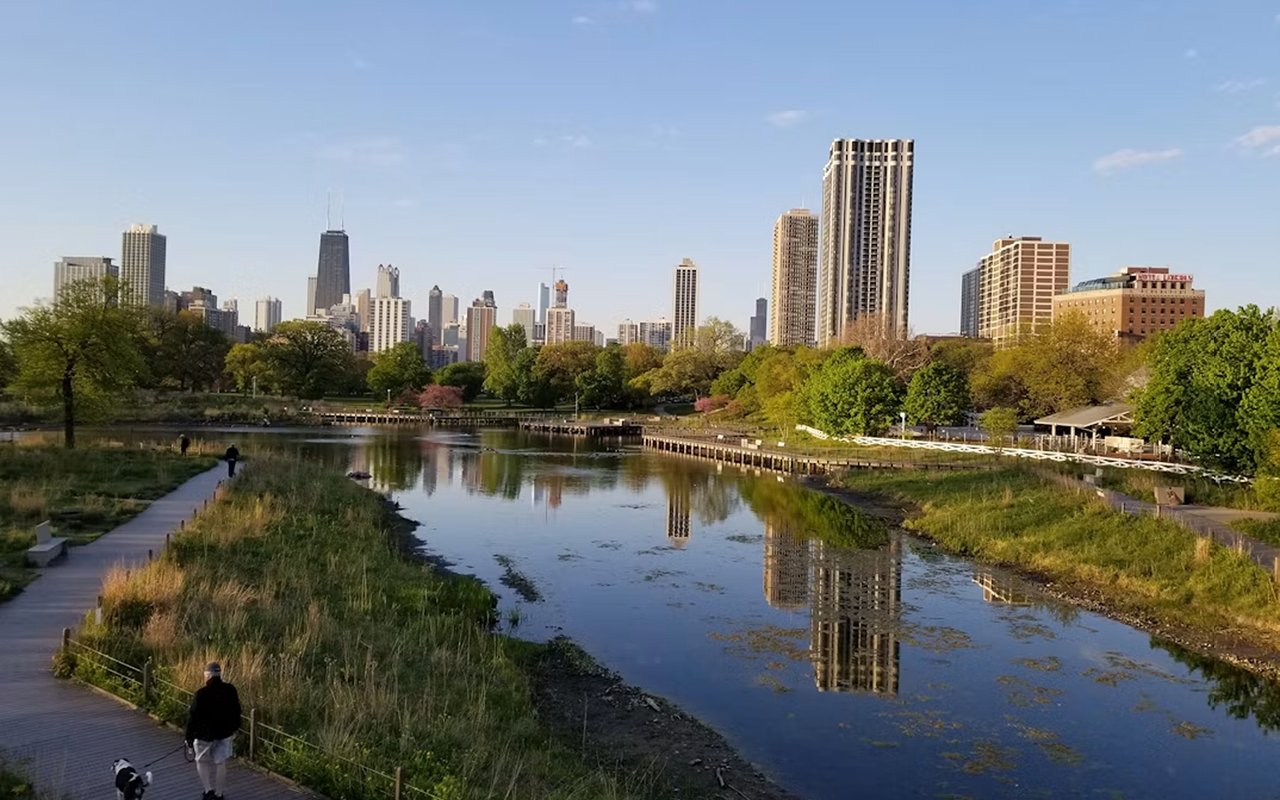 Chicago skyline reflected in a Lincoln Park pond, with a boardwalk and green spring foliage.