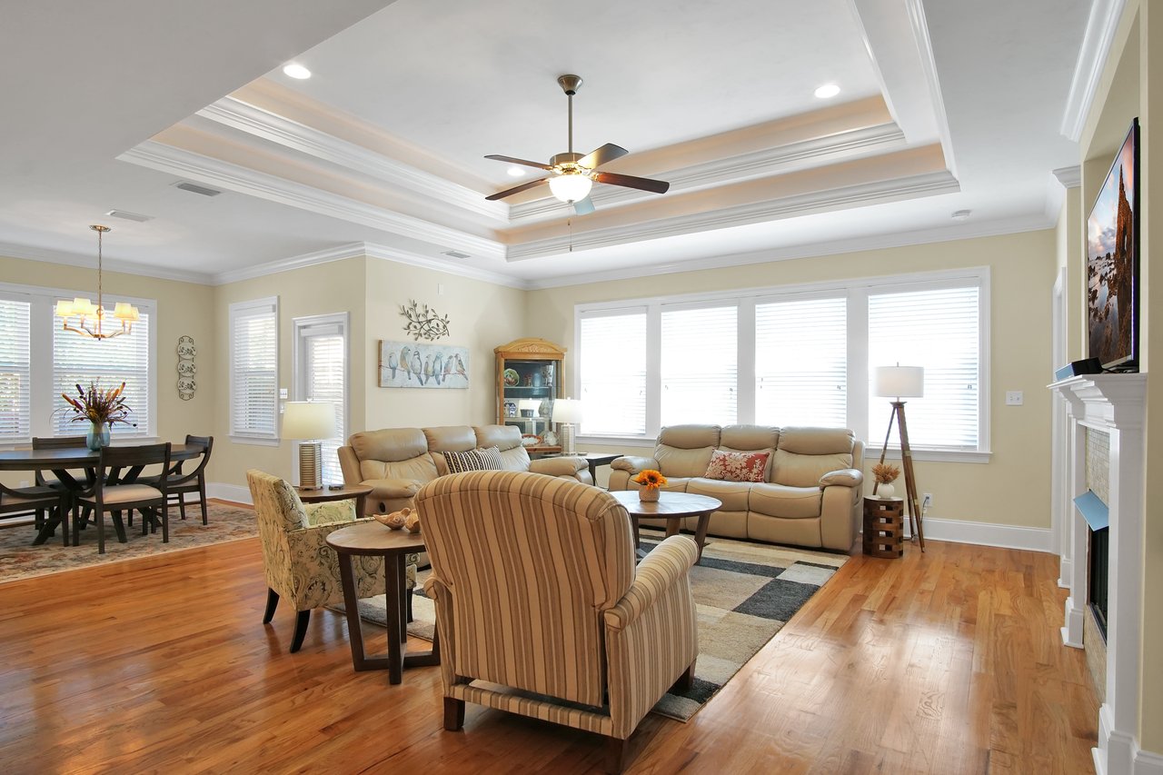 Spacious living room with beige leather sofas, striped armchairs, and wooden furniture. Bright windows, chandelier, ceiling fan, warm ambiance.