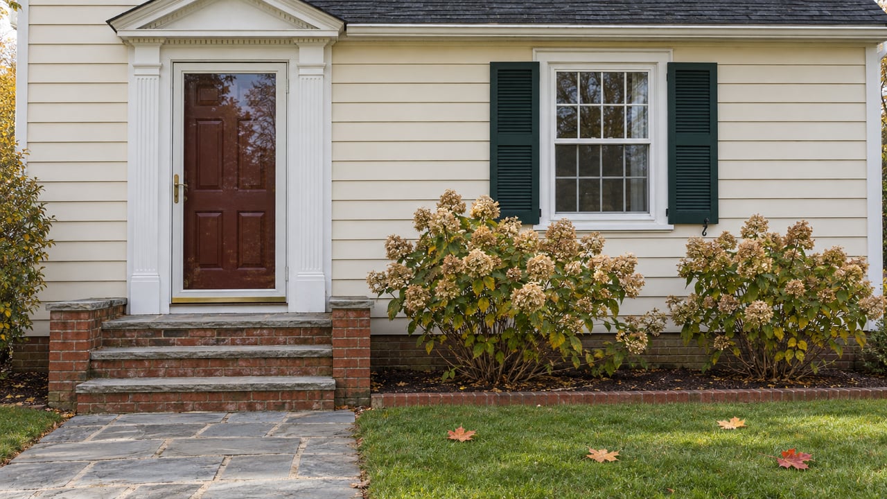 Ivory clapboard home with an oxblood entry door, worn brick steps, green shutters, and dry hydrangea flowers beside a stone walk.