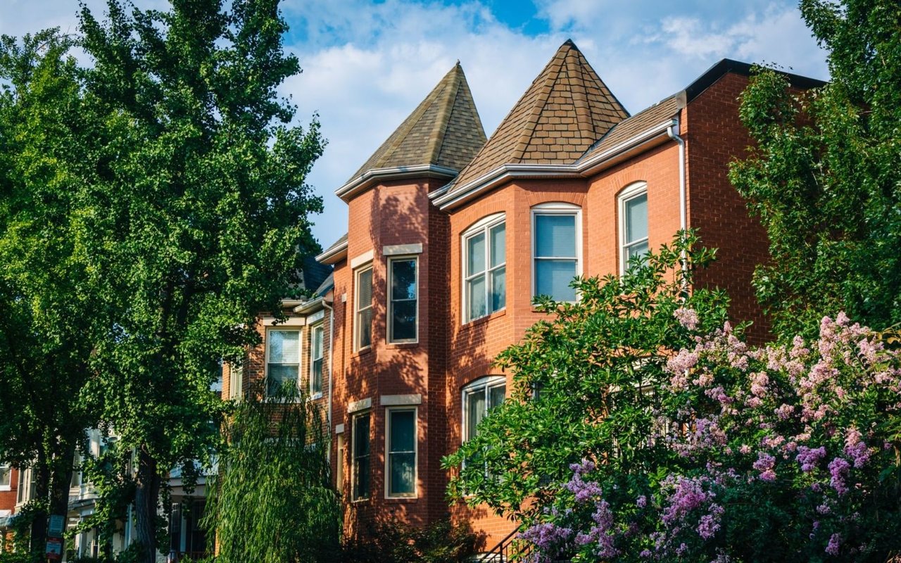 Red brick house with pointed shingle roofs, leafy trees, and flowering shrubs.
