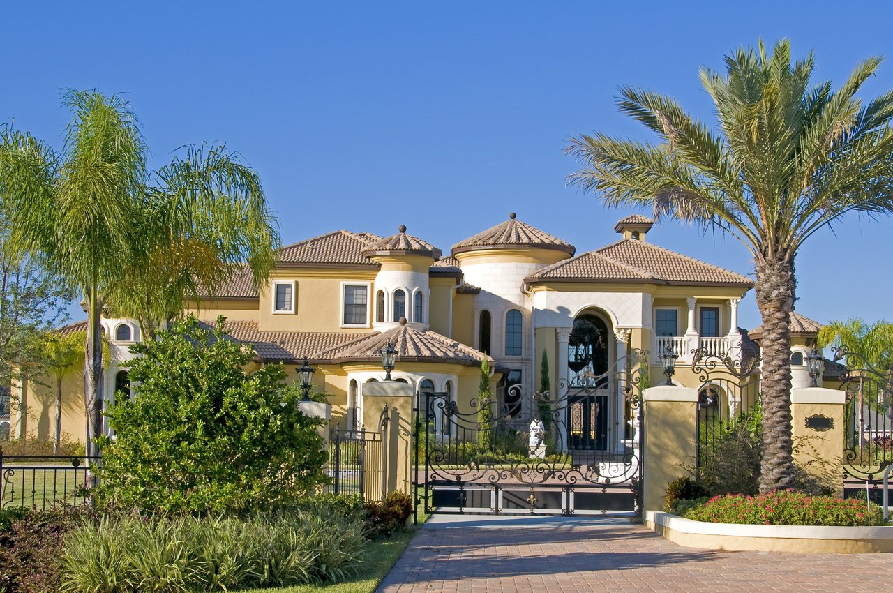 Large Mediterranean-style home behind an ornate black gate, framed by palm trees.