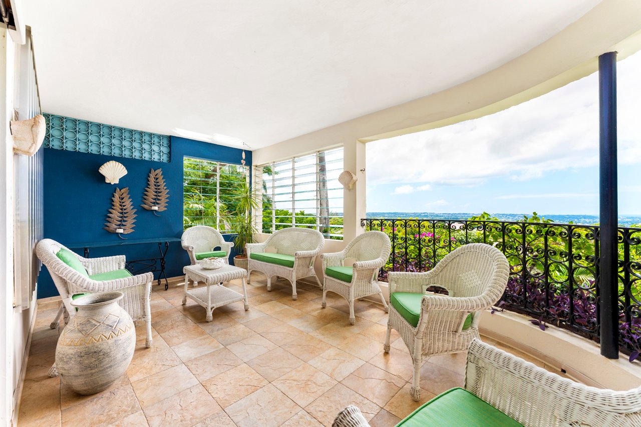 Covered terrace with wicker seating and green cushions overlooking a distant coastal horizon.