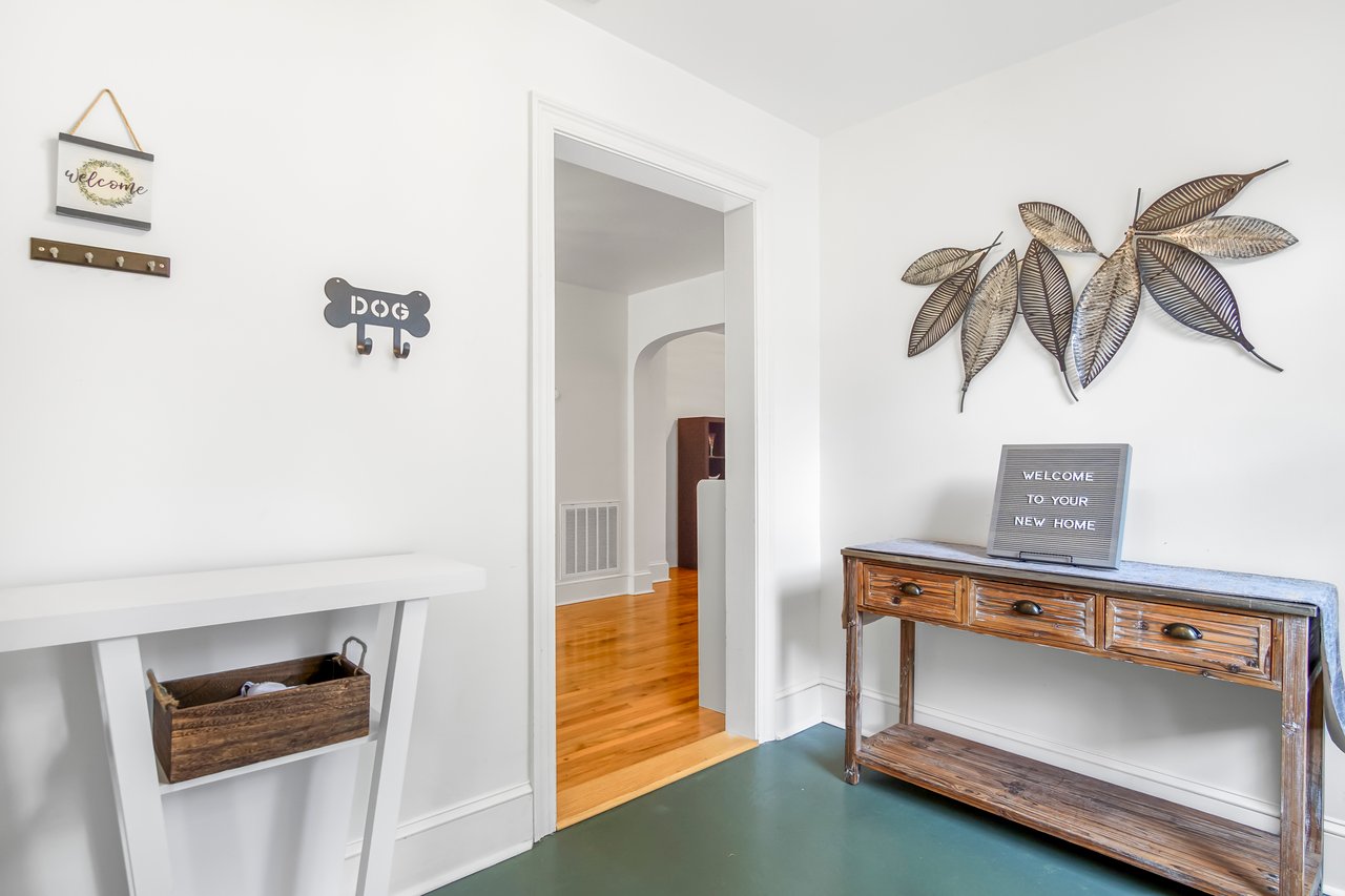 A bright hallway with wooden floor and a white wall. A rustic table on the right holds a decorative sign. Metal leaves adorn the wall above. Cozy ambiance.
