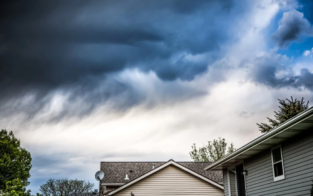 Storm clouds gather over suburban homes and leafy trees.
