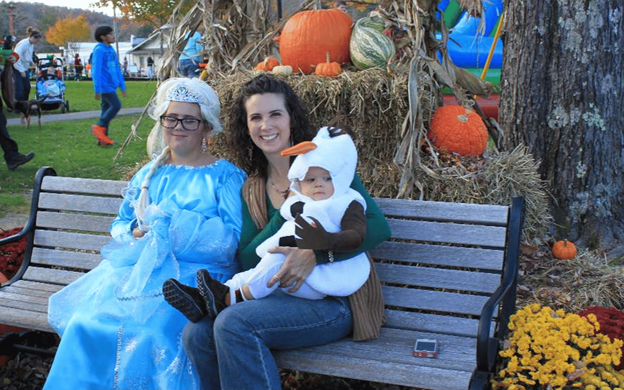 Three people in Halloween costumes sit on a bench among pumpkins and hay.