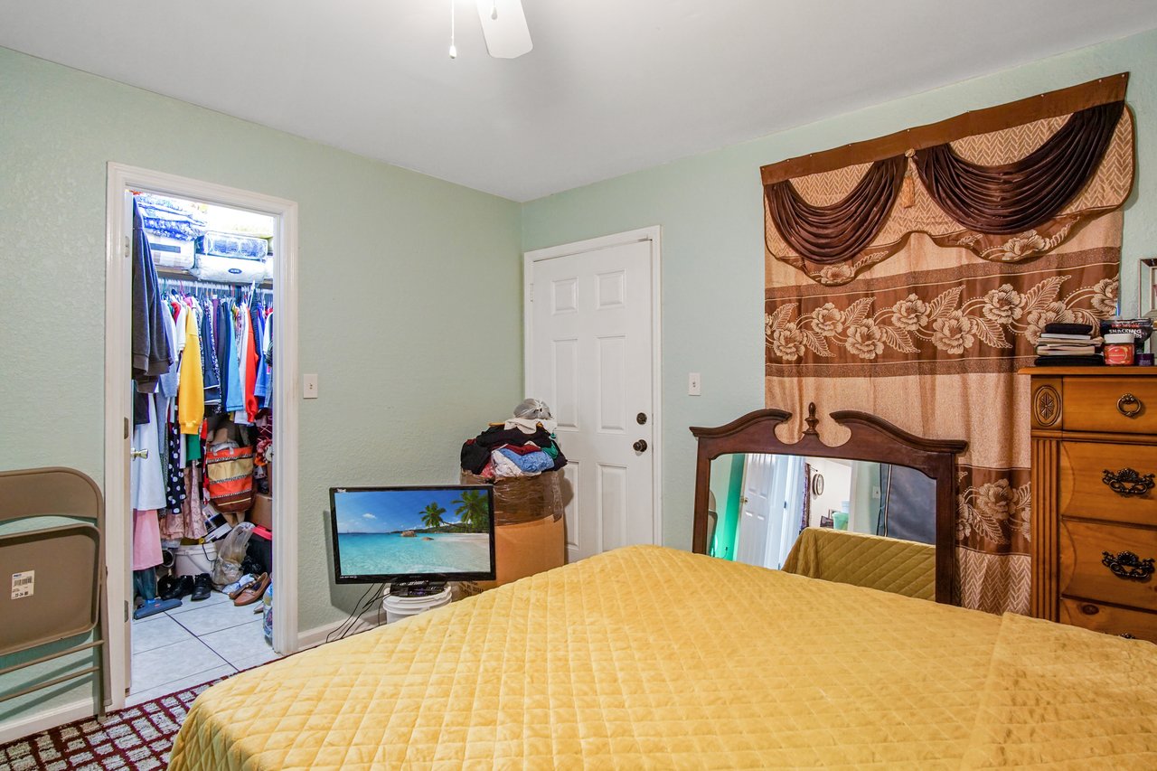 Bedroom at 2506 Nugget Lane in Tallahassee, Florida, with tile flooring and light green walls. The room includes a bed with yellow coverlet, a wooden dresser, and a decorative curtain over the window. A door leads to a walk-in closet with shelving and hanging storage.