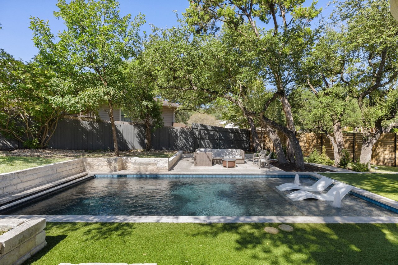 Backyard swimming pool with white loungers, stone edging, mature trees, and a fenced patio.