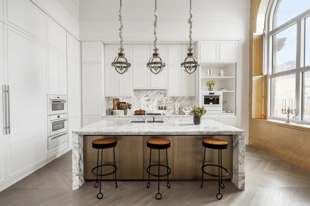 Kitchen with a marble island, white cabinetry, three pendant lights, and tall arched windows.