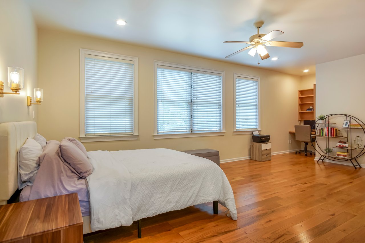 A spacious, cozy bedroom with wooden floors, a neatly made bed with white bedding, large windows, a ceiling fan, and a desk area in the corner.