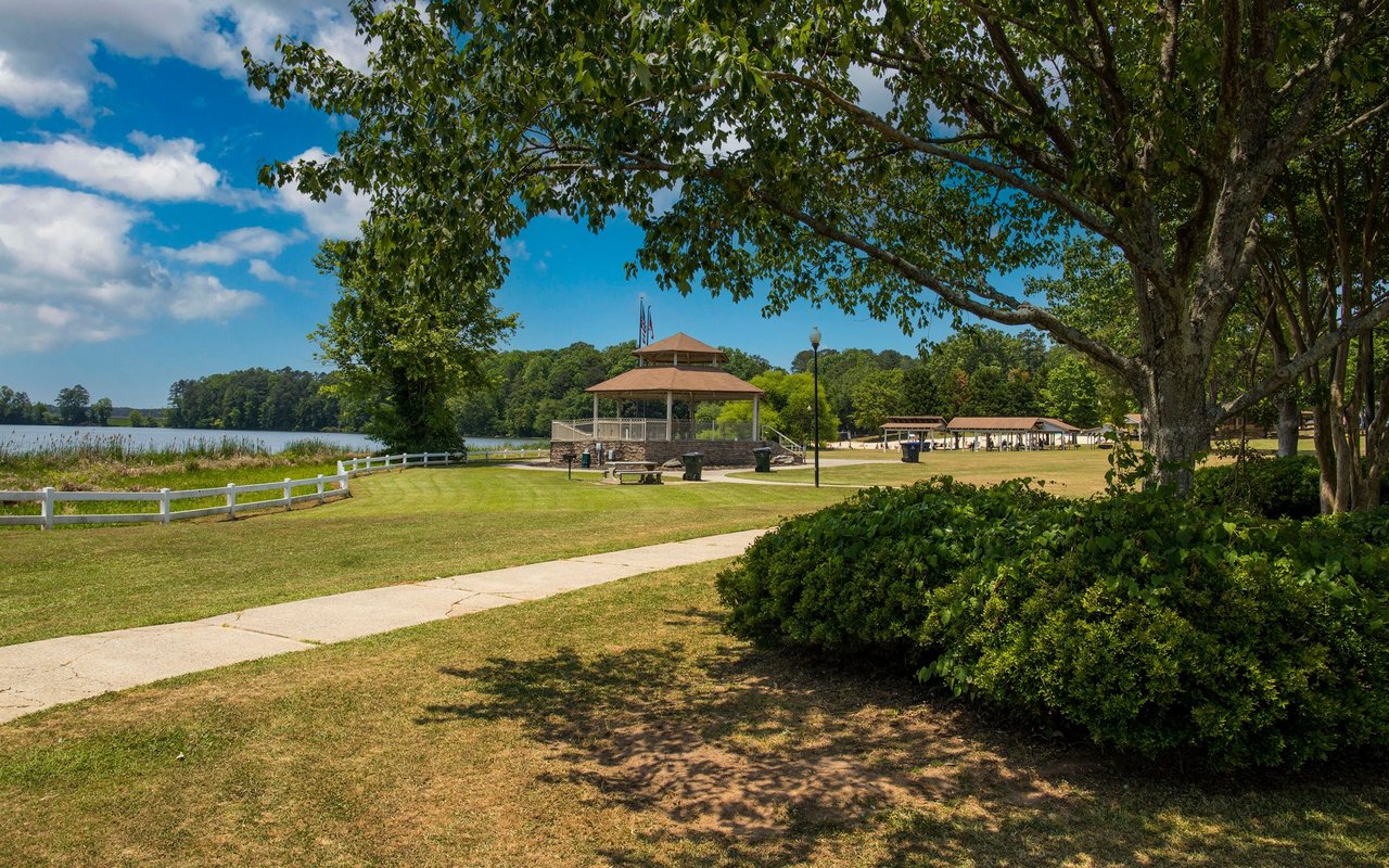 Lakeside park with a gazebo, picnic tables, and a paved path beneath mature trees.