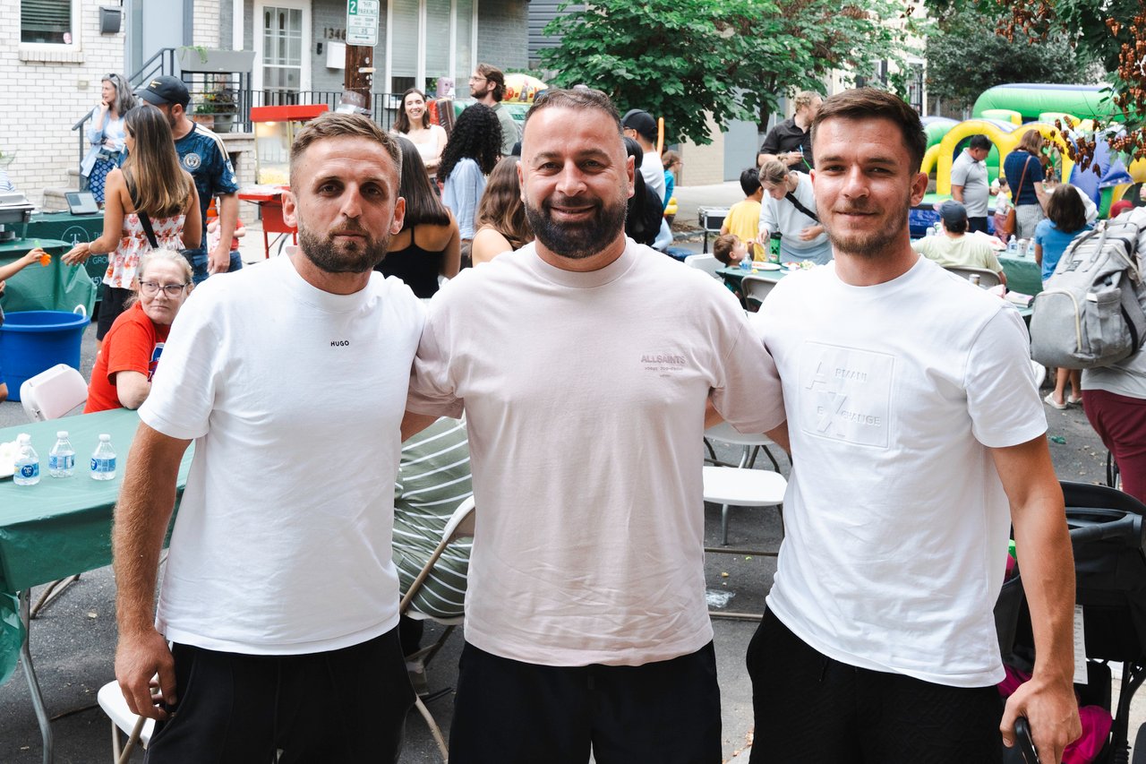 Three men pose at a neighborhood block party, with guests and a bounce house behind them.