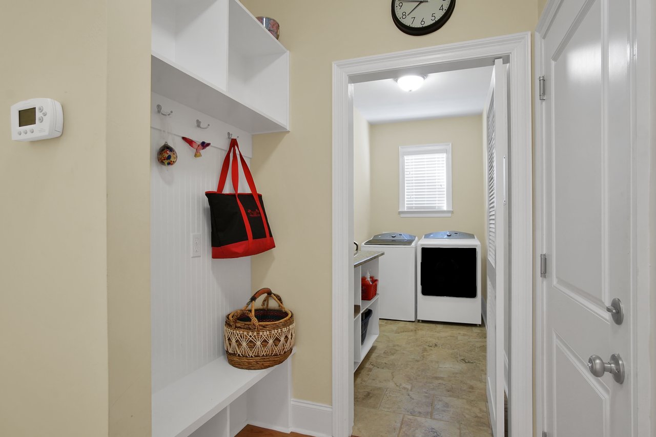 Mudroom leading to laundry room; hooks with bags, woven basket below. Washer and dryer in background, beige walls, tile floor, wall clock.