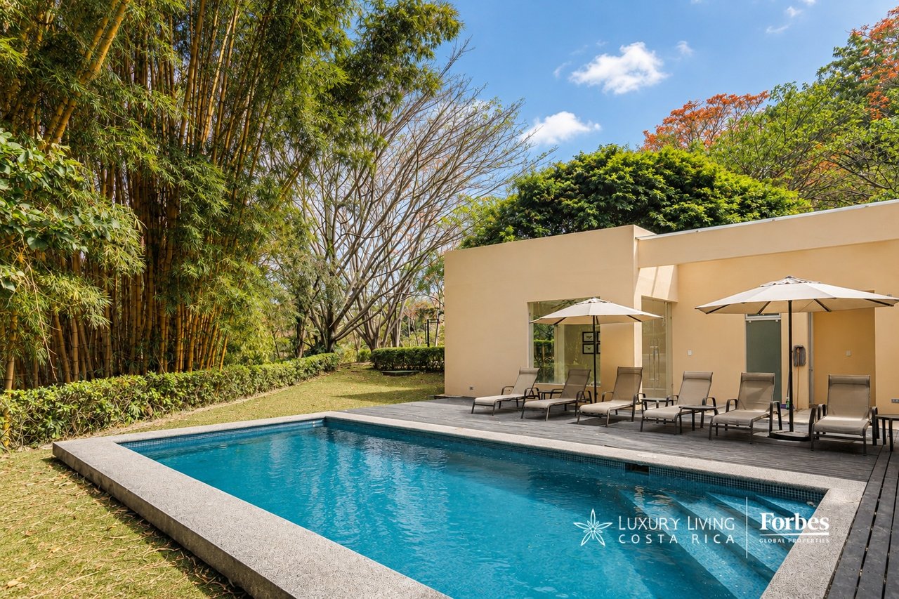 Outdoor pool beside a modern building, with a wood deck, lounge chairs, umbrellas, and bamboo landscaping.