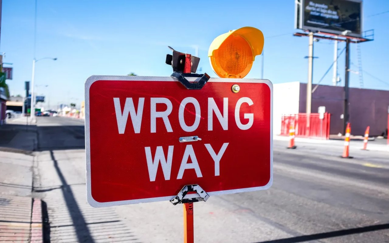 A bright red "WRONG WAY" sign is shown, with a yellow flashing light on top, placed in a street with traffic cones.