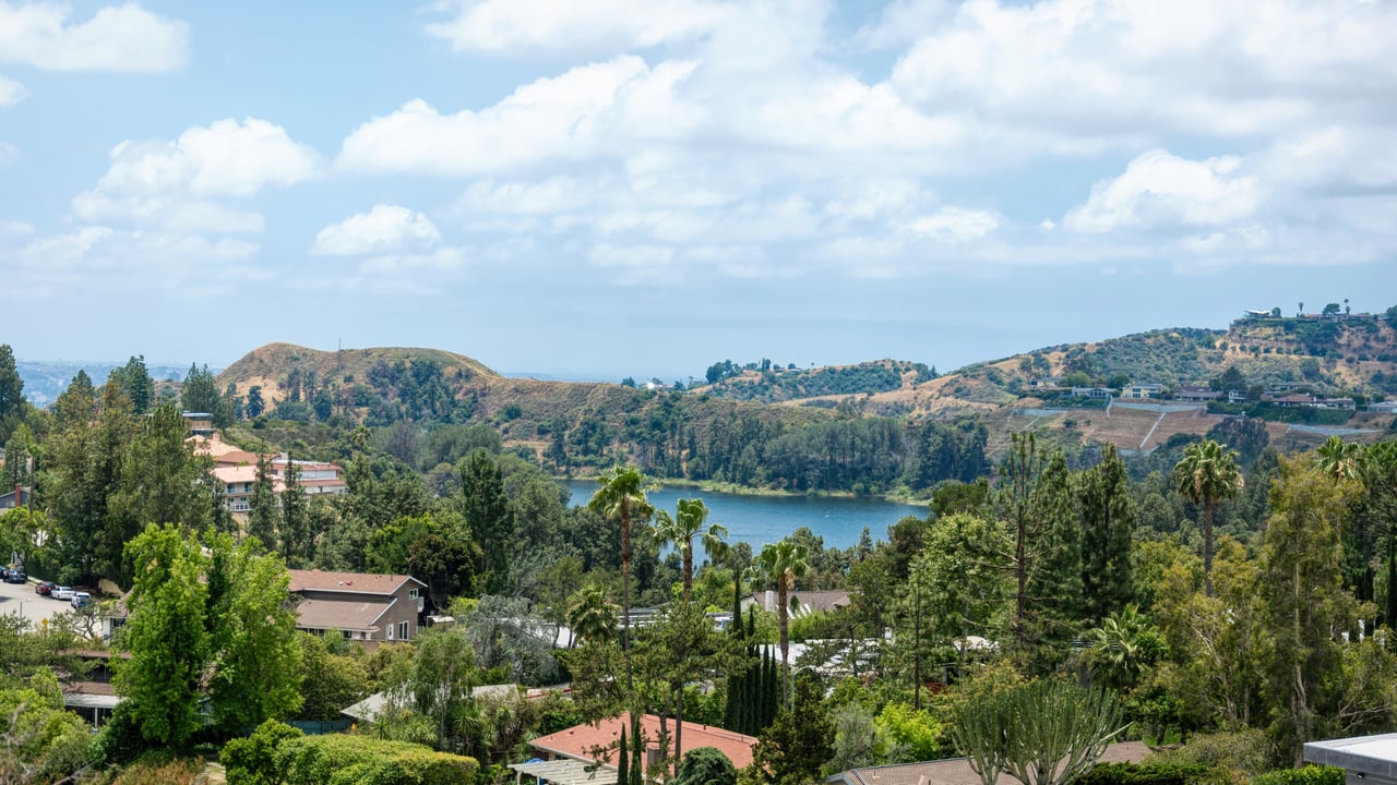 Panoramic view of a leafy hillside neighborhood, reservoir, and rolling hills beneath a partly cloudy sky.