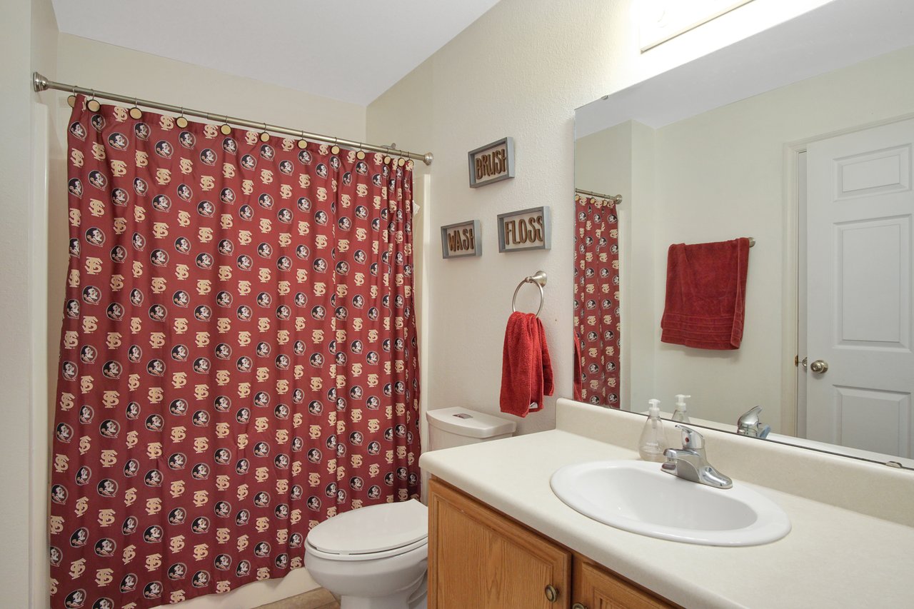 Bathroom with red themed decor, featuring a patterned shower curtain, red towel, and signs reading "Brush," "Wash," "Floss." Simple, tidy atmosphere.