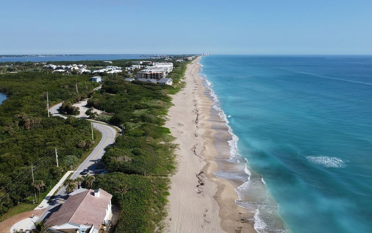 Aerial coastline with a long sandy beach, turquoise water, coastal homes, and a winding road.