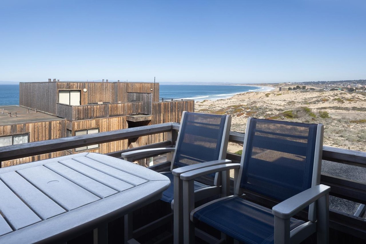 Furnished deck overlooking a wooden coastal home, sandy dunes, and ocean.