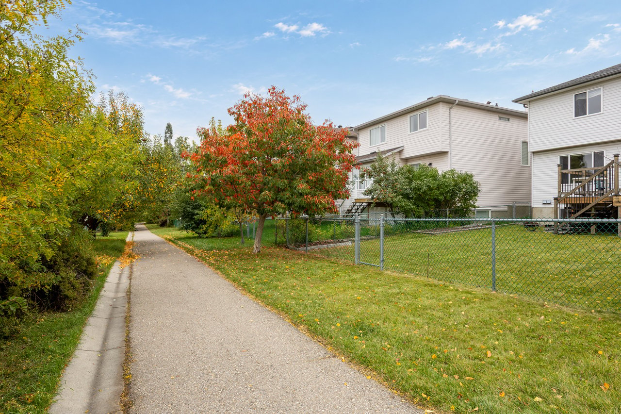 Rear exterior of light-colored homes beside a fenced lawn and tree-lined pathway.