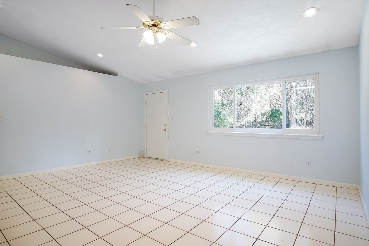 Empty room with light blue walls, white tiled floor, and a ceiling fan with lights. Large window on the right shows greenery outside. Quiet and airy atmosphere.