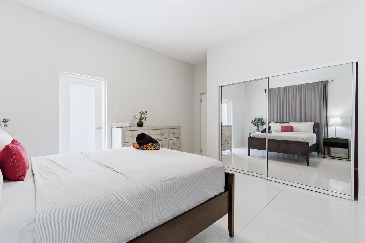 Furnished bedroom with a white bed, red pillows, and mirrored closet doors reflecting the room.