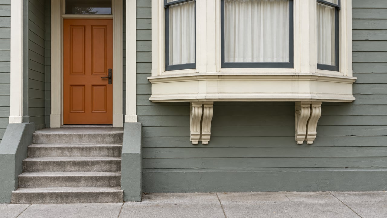 Gray-green wood facade with cream bay-window trim, a burnt-orange front door, and weathered concrete entry steps.