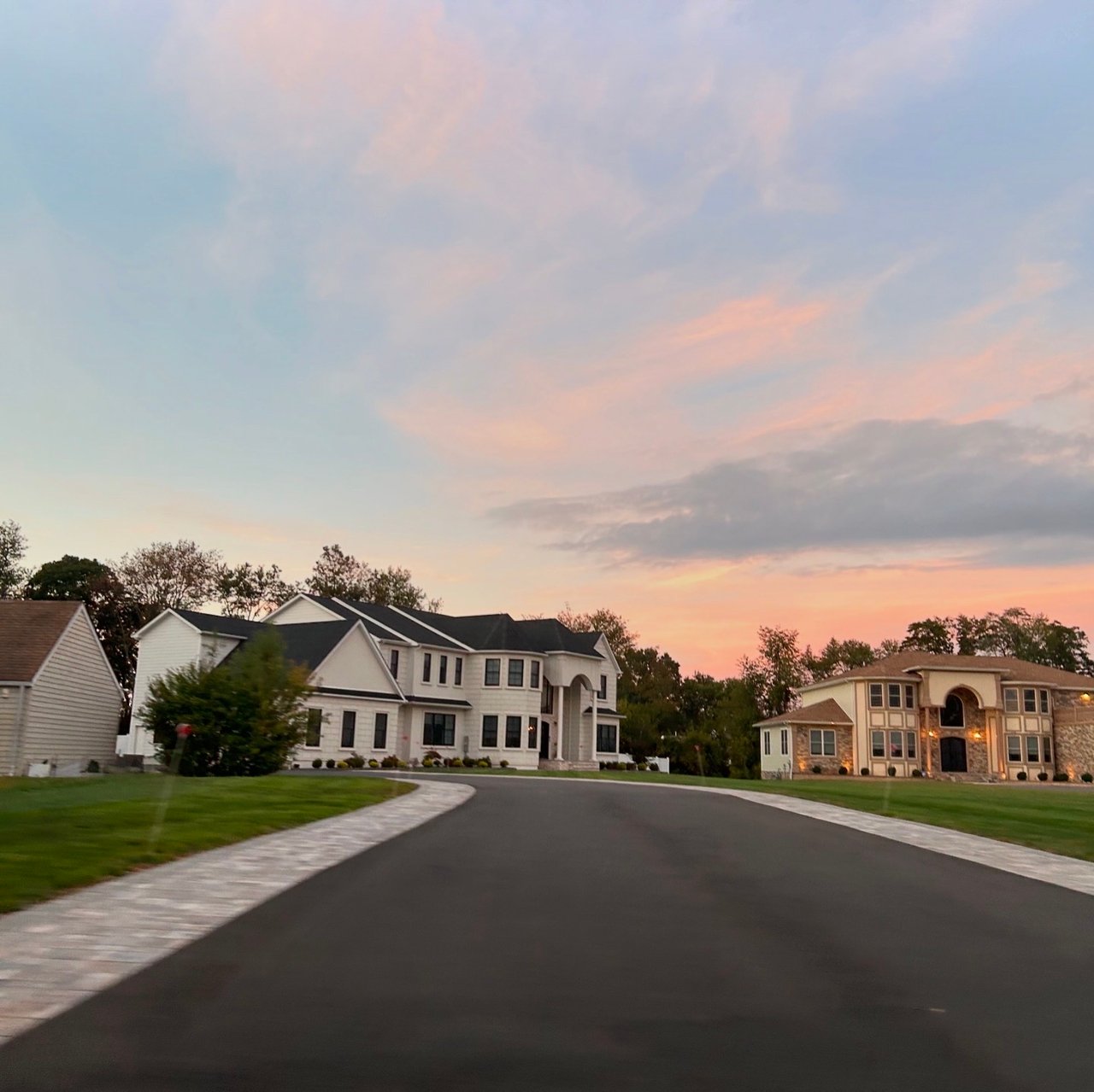 Suburban street at sunset with two large modern homes, manicured lawns, and a paved driveway under a colorful sky with pink and blue hues.