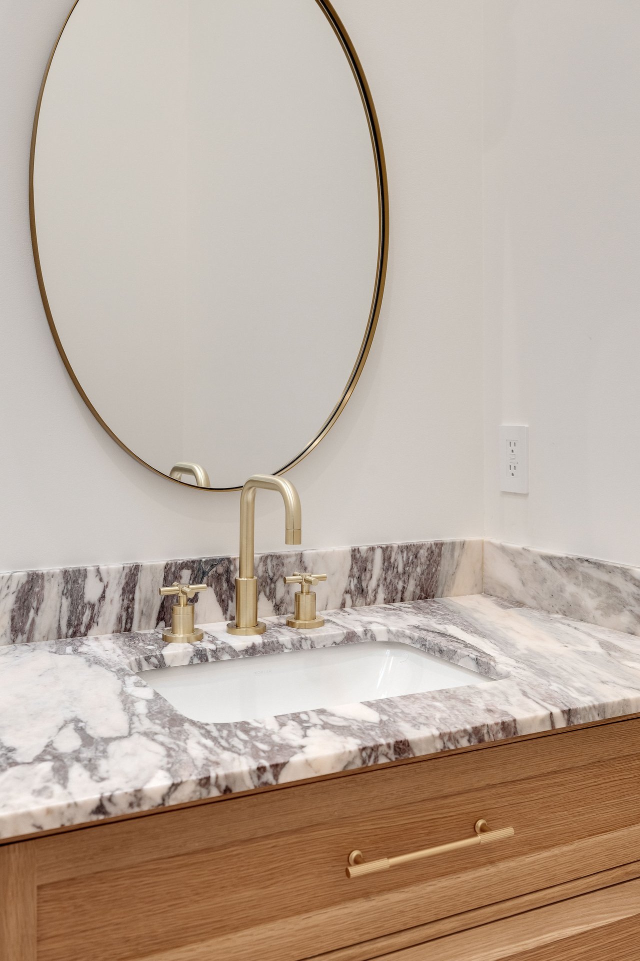 Bathroom vanity with veined marble, brass fixtures, an oval mirror, and an oak cabinet.