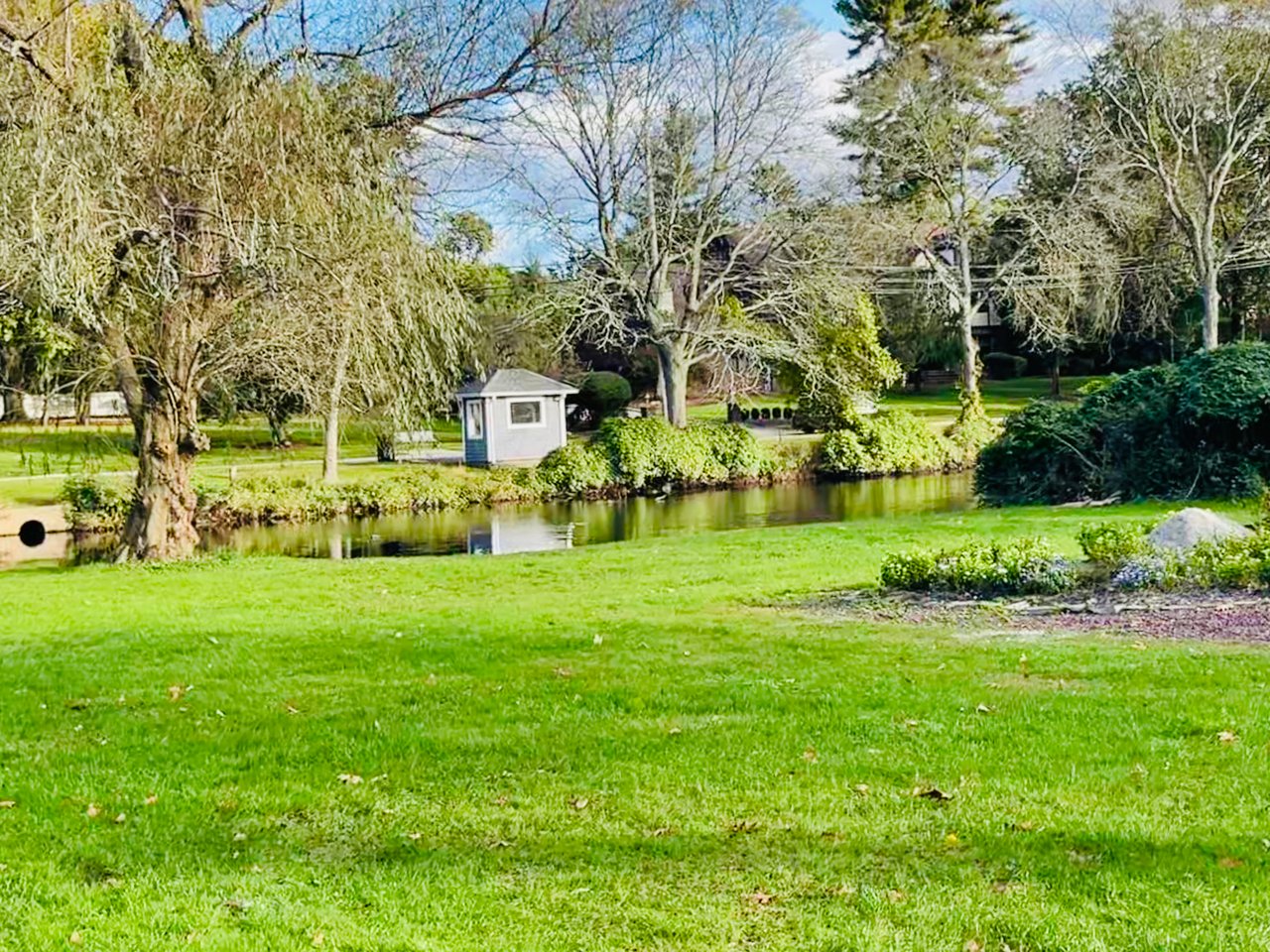 A view of the tranquil Demarest Duck Pond surrounded by bright green grass and large trees in early spring.