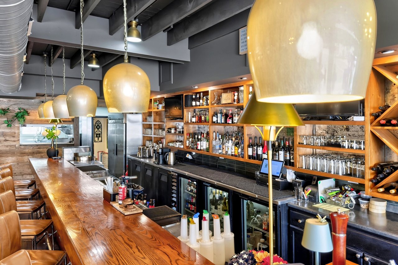 Restaurant bar with a polished wood counter, leather stools, and bottle-lined shelves.