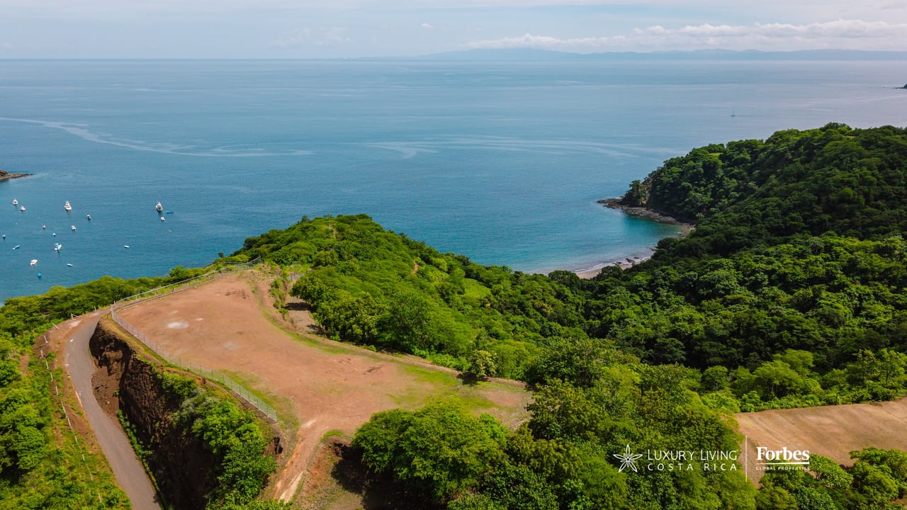 Cleared coastal residential lot overlooking a blue bay, with a winding road and forested hillsides.