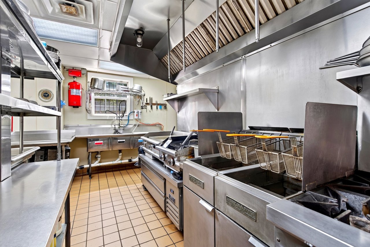 Commercial restaurant kitchen with stainless-steel cooking equipment, fryers, and an overhead ventilation hood.