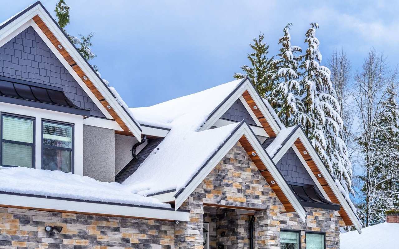 Modern house with snow-covered gabled roofs, gray shingles, and a stone facade.