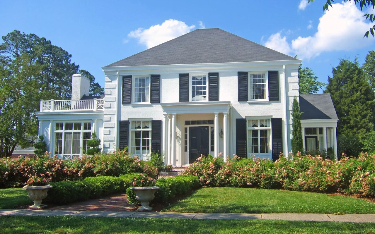 White two-story house with black shutters, a columned entrance, and flowering front gardens.
