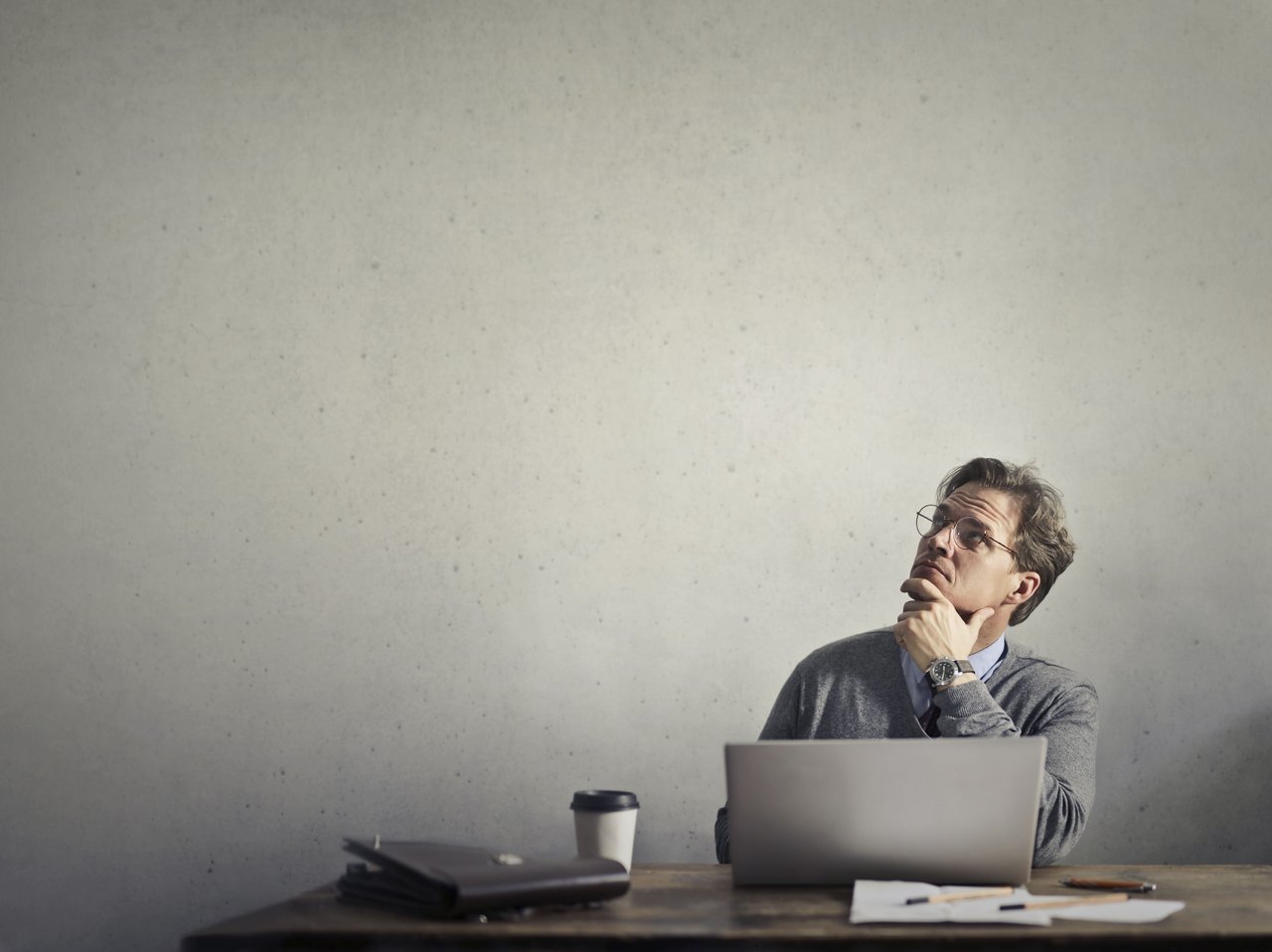 Thoughtful man with glasses at a desk, looking upward beside his laptop.