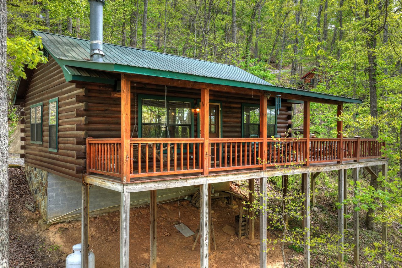 Raised log cabin with a long covered porch and green metal roof in a leafy woodland.