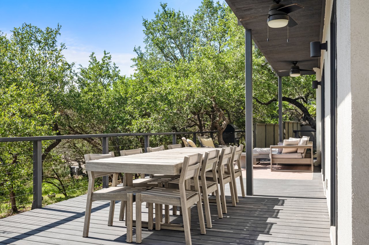 Outdoor deck with dining table and chairs, covered lounge seating, and leafy trees beyond the railing.