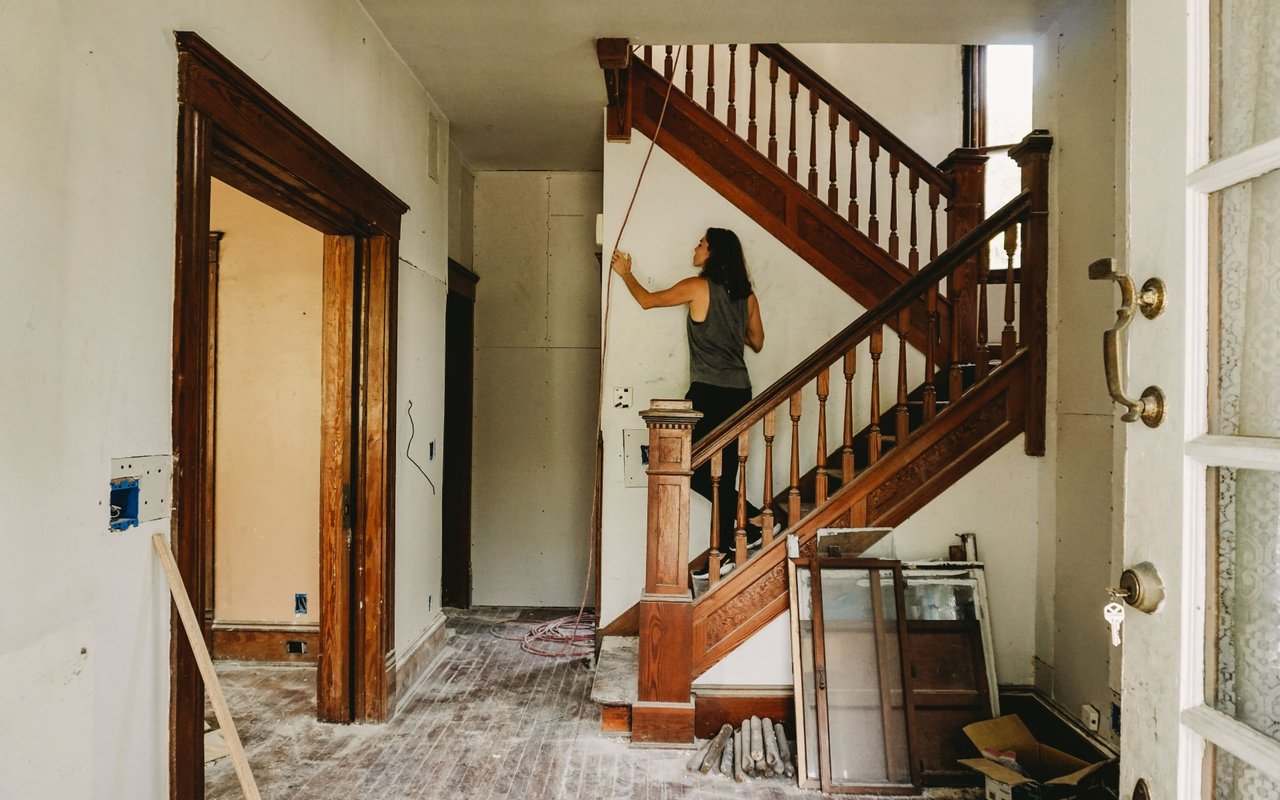 Unfinished entry hall with an ornate wooden staircase and a woman working on the wall.