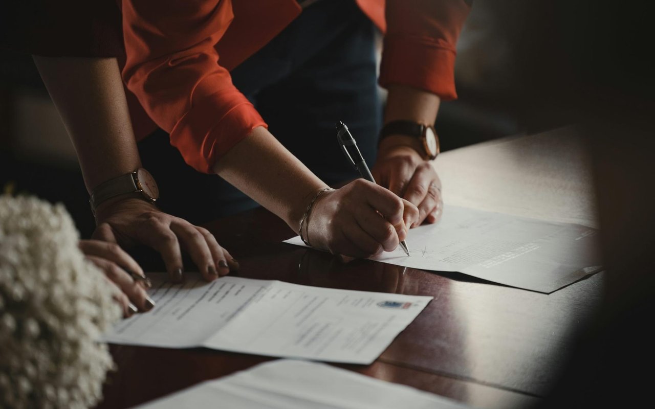 A close-up shot of two people's hands on a dark wooden table, focusing on one person signing a document.