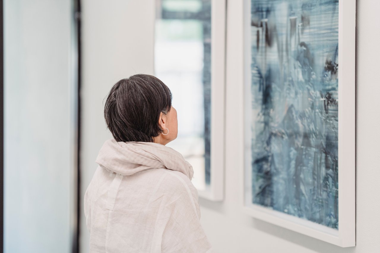 A person in a light scarf looks at framed blue abstract artwork.