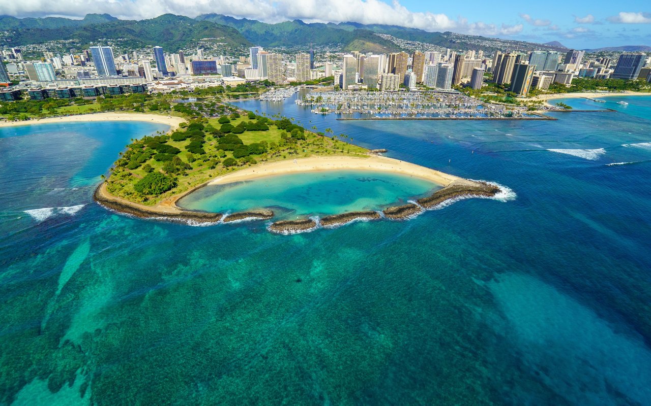 Aerial view of Honolulu’s turquoise coastline, marina, high-rise skyline, and green mountain range.