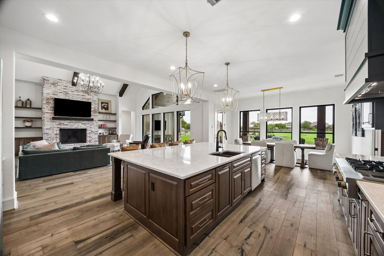 Open-plan kitchen and living area with a large island, brick fireplace, and windows overlooking a lawn.