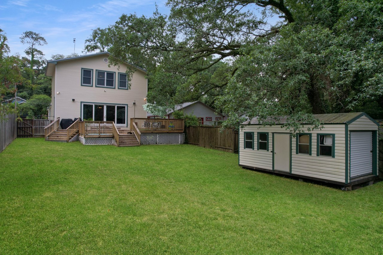A two-story house with a wooden deck overlooks a spacious green backyard. A small shed is on the right, surrounded by lush trees under a cloudy sky.