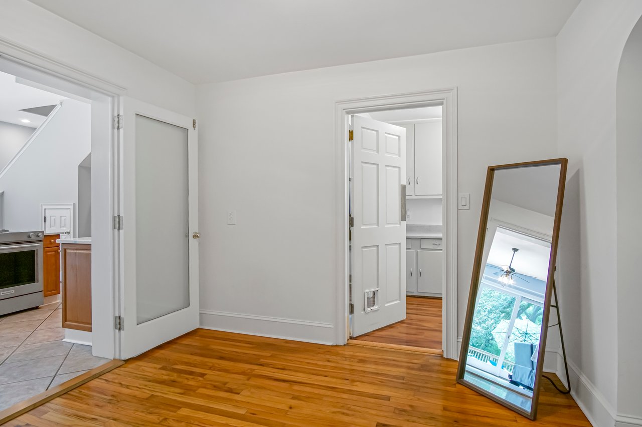 Minimalist room with wooden floors, white walls, and a large mirror reflecting a bright room. Open door to a bathroom, and a glimpse of a kitchen.