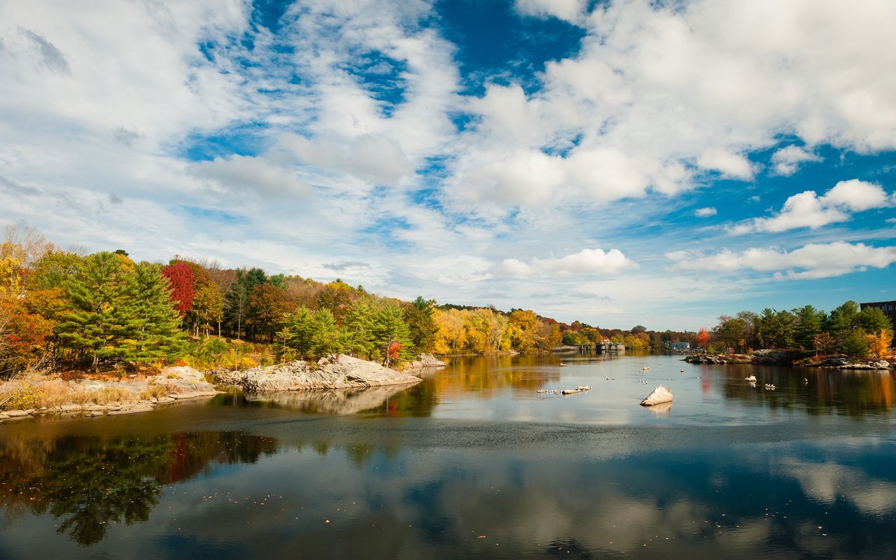 River bordered by rocky banks and colorful autumn trees under a partly cloudy sky.