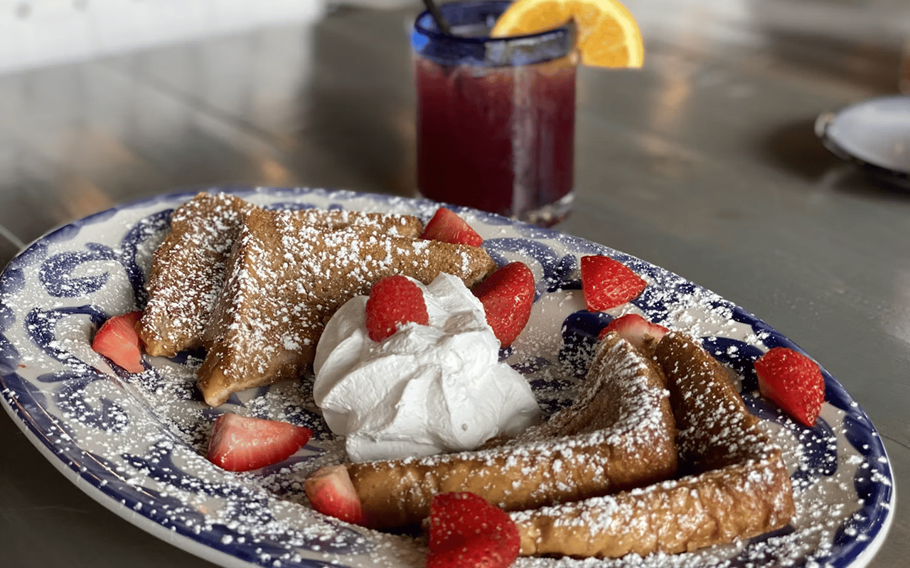 Powdered-sugar French toast with strawberries and whipped cream beside a dark red drink with orange garnish.