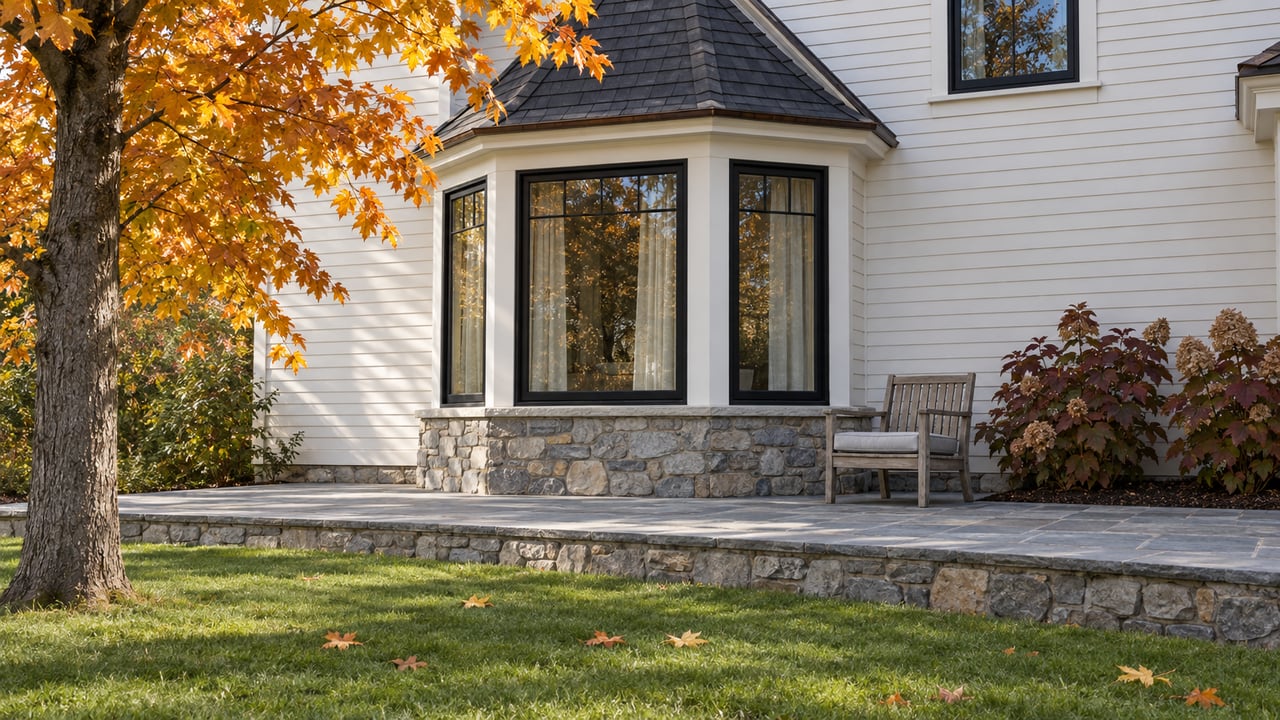 White clapboard home with black bay windows, a raised bluestone terrace, and orange autumn maple foliage beside a broad lawn.