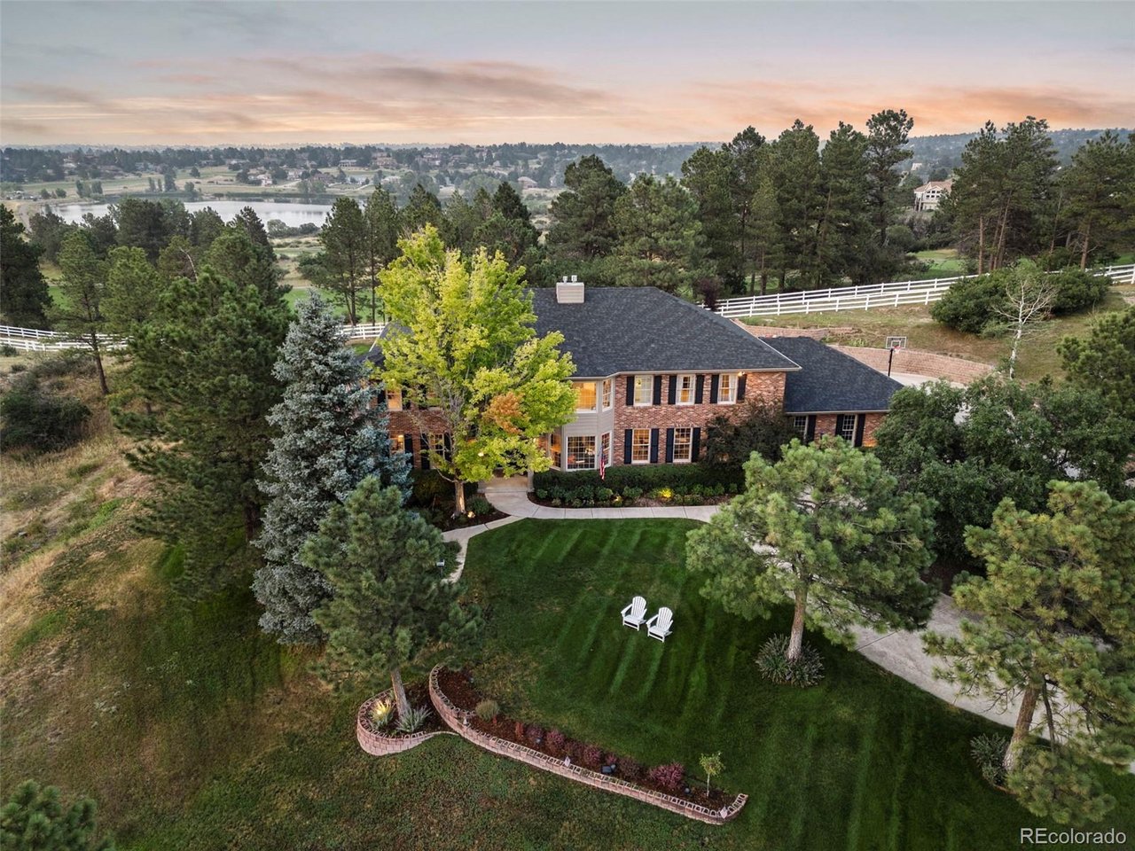 Aerial view of a brick home with landscaped grounds, a white fence, and a lake in the distance.