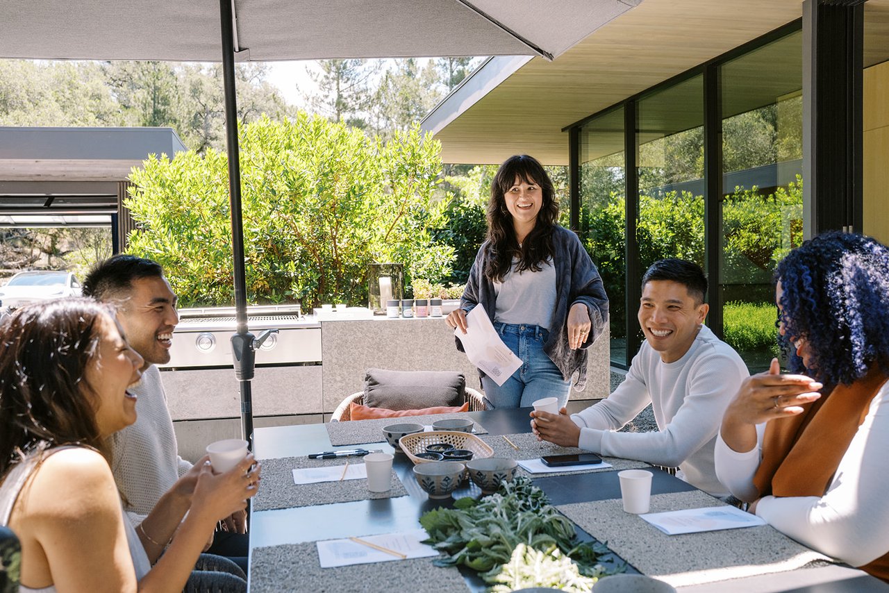 Colleagues gather around a patio table as a woman stands and speaks with papers in hand.